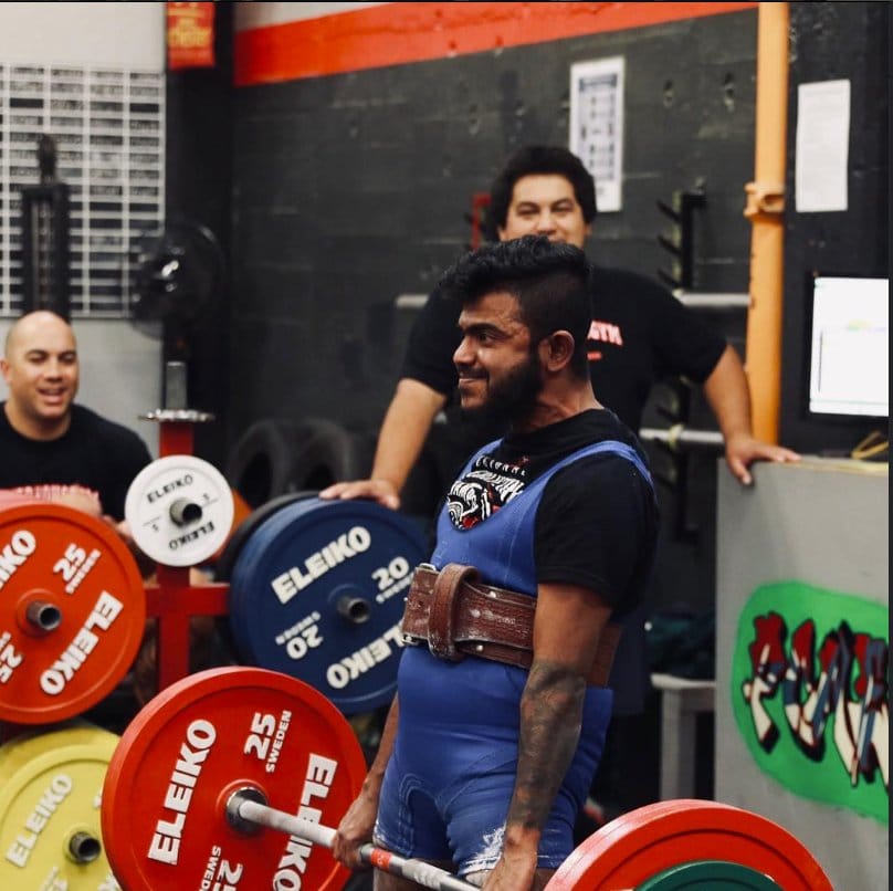 Chad Karannagoda in a lifting singlet and belt, standing behind a loaded barbell